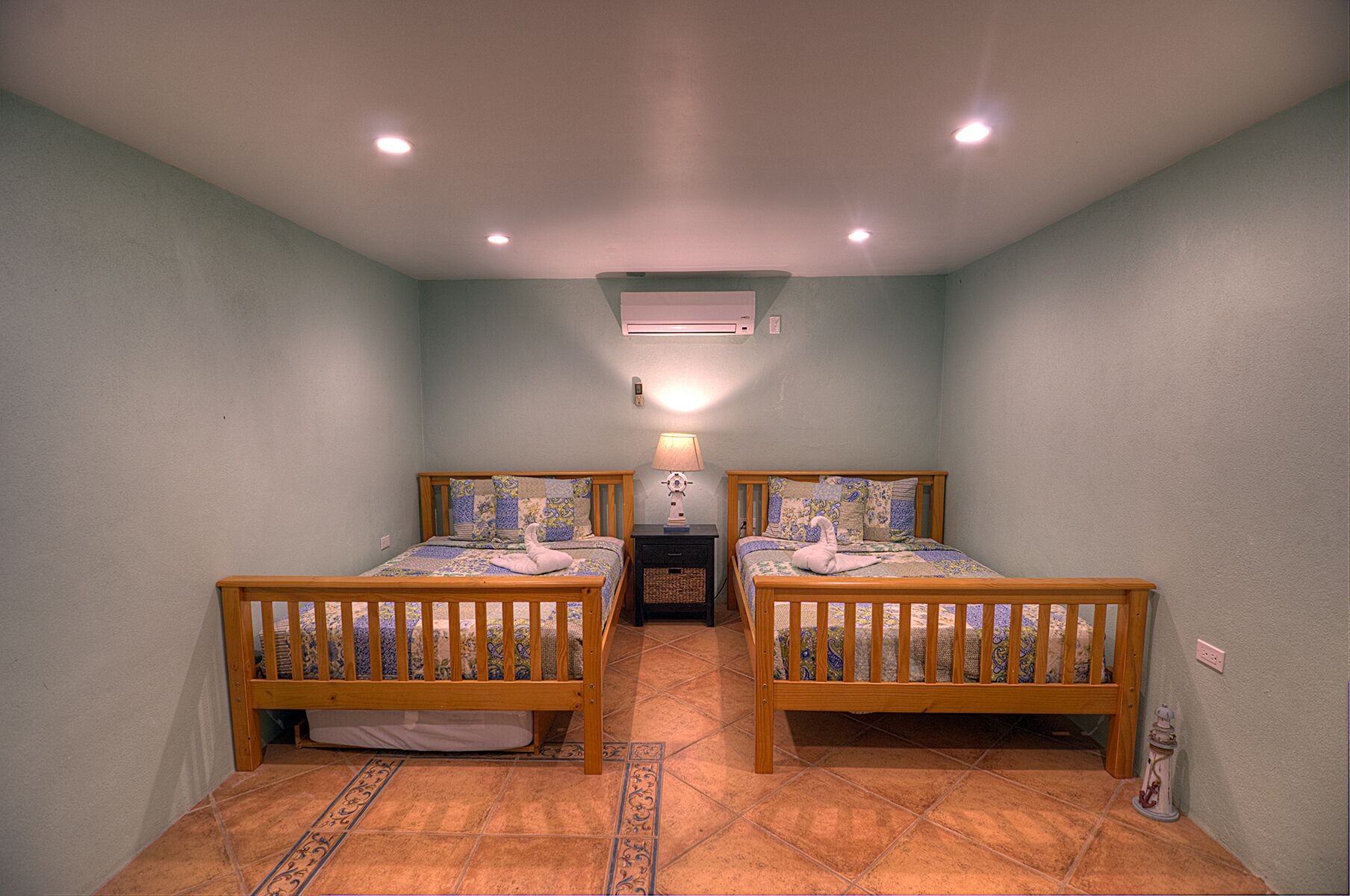 Una habitación pequeña y bien iluminada, con dos camas de madera, una mesita de noche y un aire acondicionado instalado en la pared.