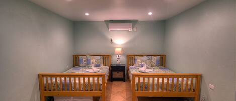 Una habitación pequeña y bien iluminada, con dos camas de madera, una mesita de noche y un aire acondicionado instalado en la pared.
