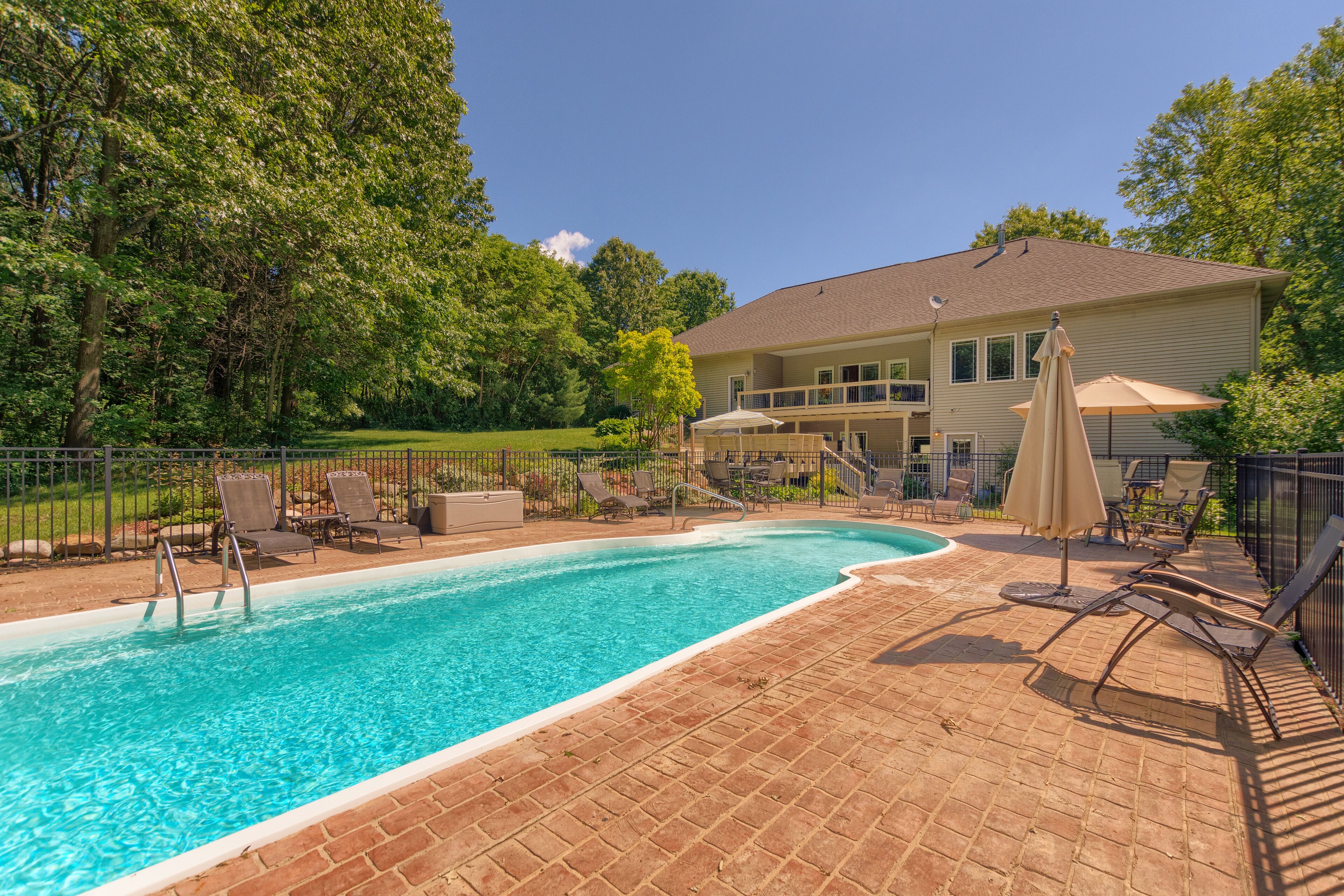 A swimming pool with lounge chairs and umbrellas, surrounded by a brick patio and a house with a balcony.