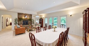A spacious living room with a fireplace, a dining area with a table and chairs, and a view of the outdoors through large windows.