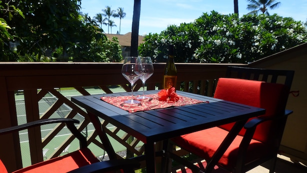 A table with a bottle and glasses on a red mat, surrounded by red chairs on a balcony with a view of palm trees and a tennis court.