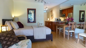 A bedroom with a bed, bedside table, lamp, and a view into the kitchen.