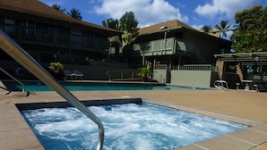 A hot tub area with a pool, lounge chairs, and a building with a balcony in the background.
