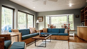 Un salón espacioso con un sofá azul, una mesa de centro de madera y amplias ventanas que ofrecen vistas al exterior.