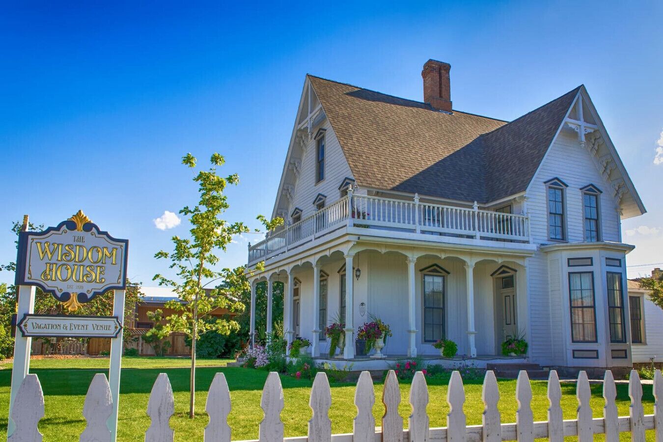 Une maison à deux étages avec un porche, une enseigne indiquant qu’il s’agit d’un lieu de location pour les vacances et d’un espace événementiel, et une clôture blanche en piquets.