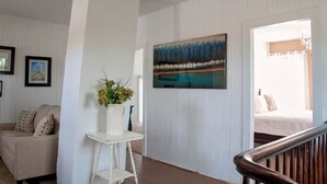 A room with a bed, a white wall with a painting, a small table with a vase of flowers, and a staircase.
