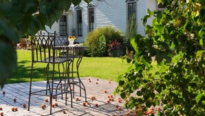 A patio with a metal table and chairs, surrounded by a garden with fruit trees and a white house in the background.