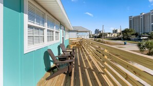 Une terrasse en bois avec des chaises, un bâtiment aux murs turquoise et aux bordures blanches, et un ciel bleu dégagé.