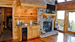 Interior de una cabaña de troncos con chimenea de piedra, armarios de madera y un televisor montado encima.