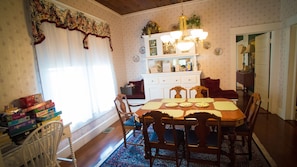 Une salle à manger avec une table en bois, des chaises et un lustre. Il y a une fenêtre avec des rideaux et un lambrequin à motifs floraux. Un buffet blanc aux portes vitrées est adossé au mur.