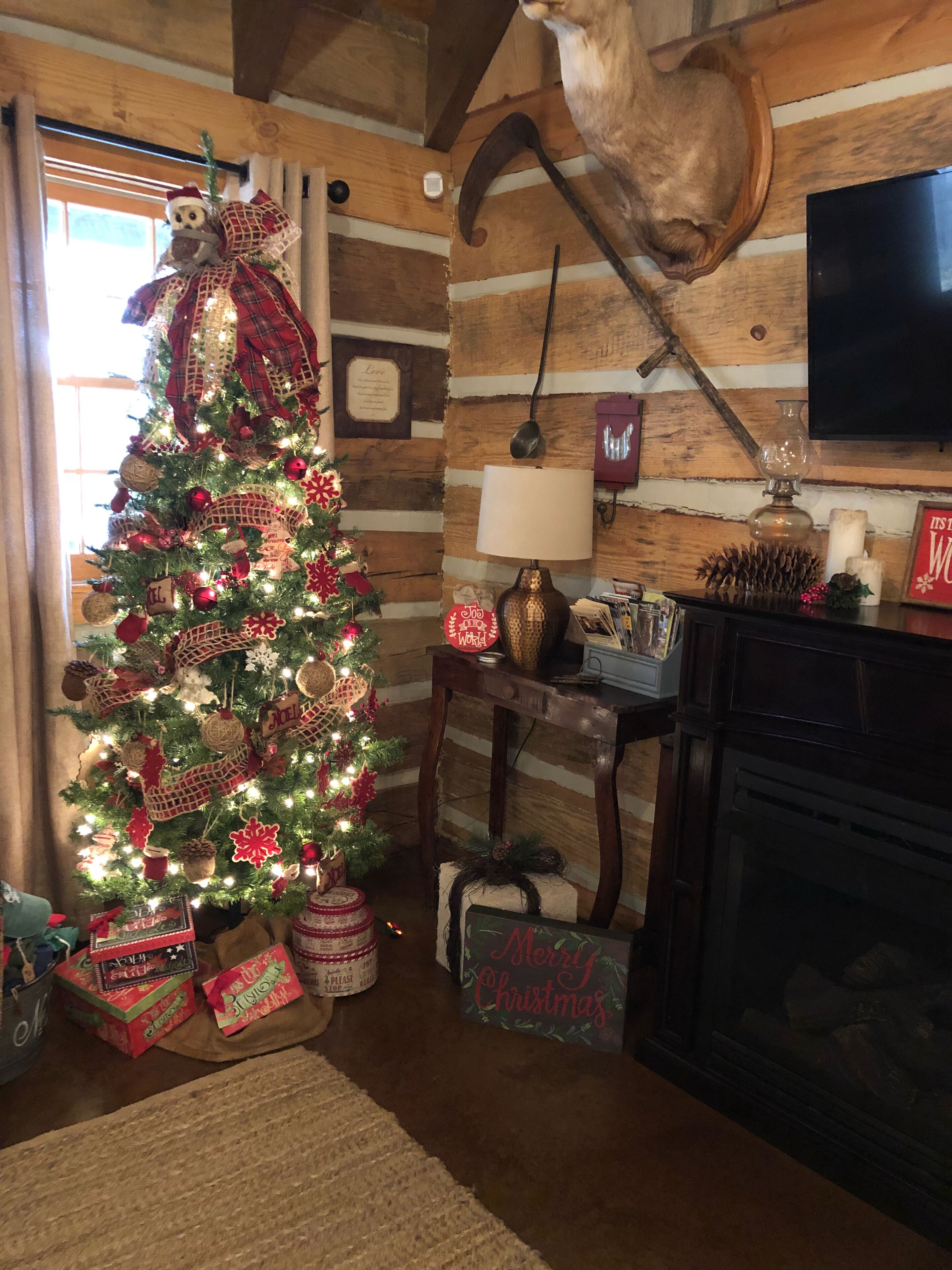A cozy cabin interior with a decorated Christmas tree, wooden walls, a fireplace, and a mounted cat.