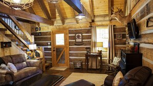 A log cabin interior with a wooden staircase, leather sofas, a fireplace, and a mounted deer head.
