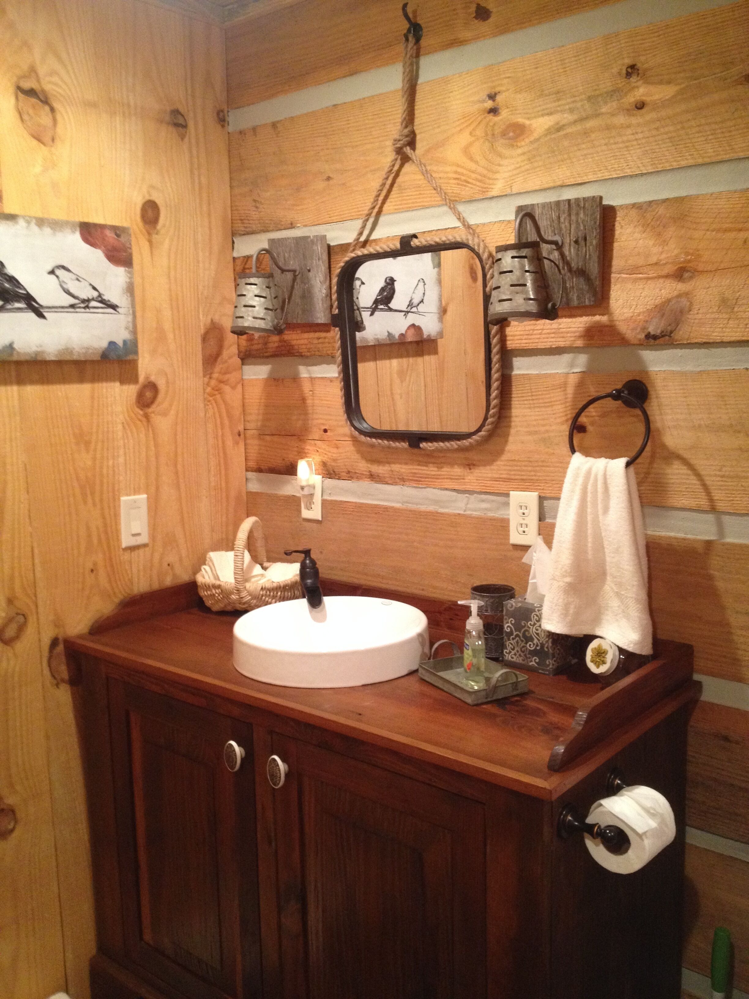 A wooden bathroom with a round sink, a hanging mirror, a towel rack, and a framed picture of birds.