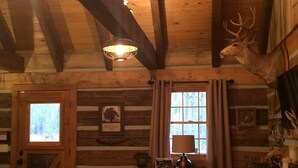 A log cabin interior with a wooden ceiling, a deer head decoration, a window with curtains, a door with a view, and a table with a lamp and a framed picture.