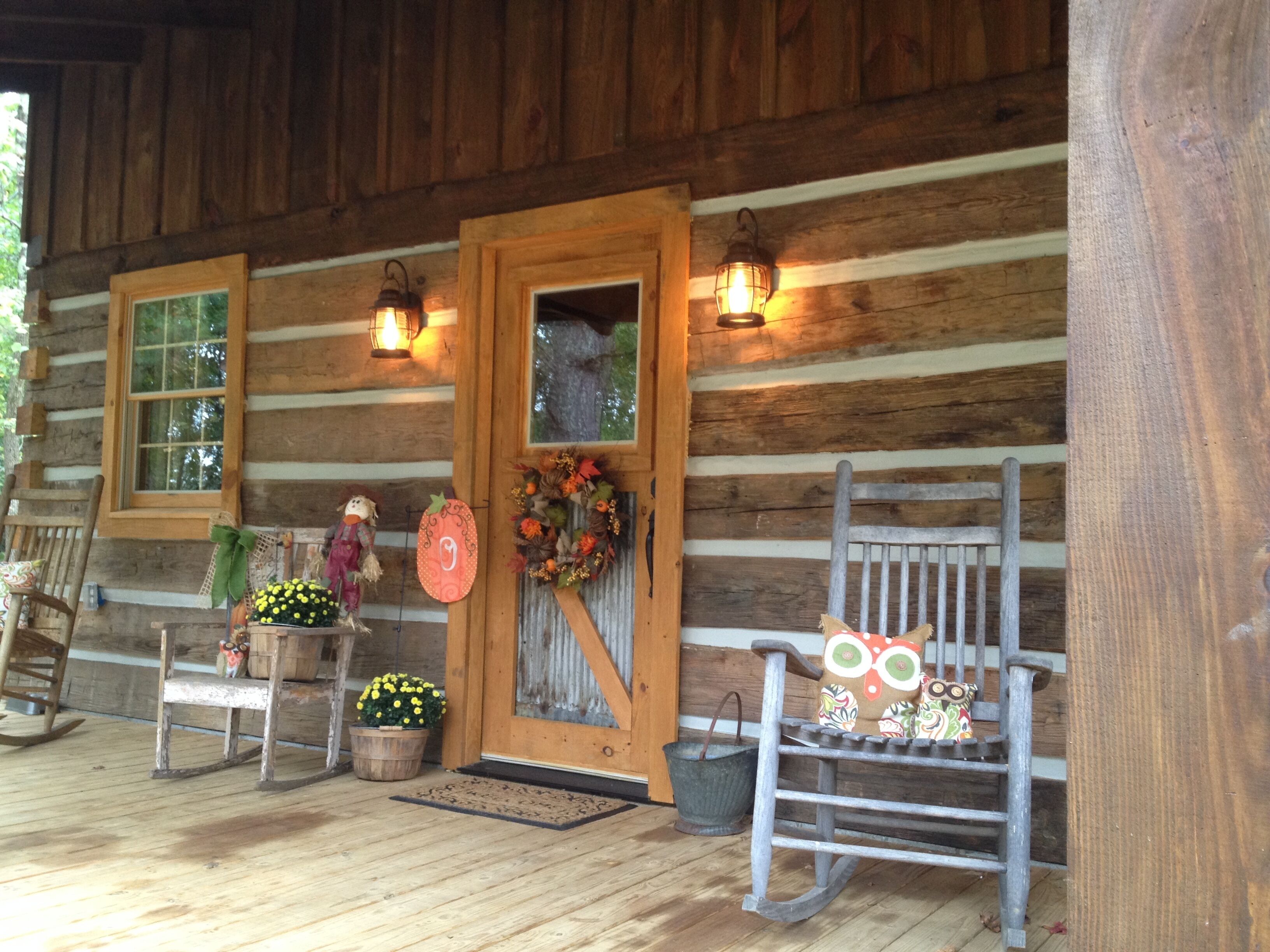 A rustic wooden cabin with a porch, a rocking chair, a door with a wreath, and two windows with lanterns.