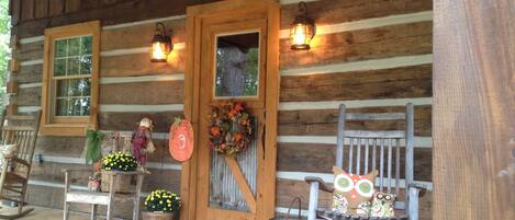 A rustic wooden cabin with a porch, a rocking chair, a door with a wreath, and two windows with lanterns.