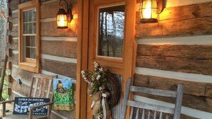 A rustic wooden cabin with a porch, two wall-mounted lanterns, a welcome sign, a bench, and a bucket.