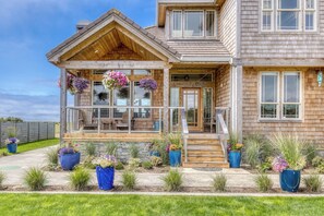 A two-story wooden house with a porch, stone foundation, and potted plants.