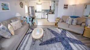 A living room with a sofa, a coffee table, and a rug. There is a kitchen in the background with a refrigerator and a sink.