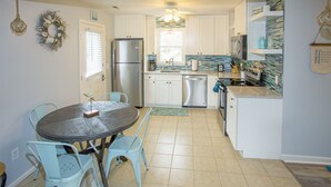 A compact kitchen with white cabinets, a stainless steel refrigerator, a dishwasher, and a dining area with a round table and blue chairs.