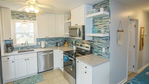 A modern kitchen with white cabinets, a stainless steel dishwasher, a built-in oven, and a microwave. There is a window above the sink and a ceiling fan.