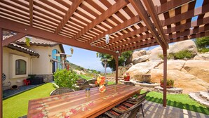 An outdoor patio with a wooden table and wicker chairs, a pergola overhead, and a view of a house with a stone facade and a tiled roof.