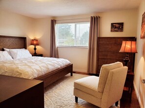 A hotel room with a bed, a chair, a nightstand with a lamp, a window with curtains, and a wooden headboard. - Riverside Beach House (Seaside)