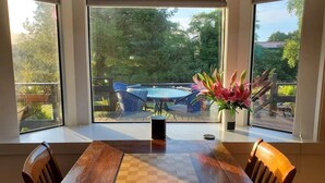 Uma sala de jantar com uma mesa de madeira, cadeiras e um tabuleiro de xadrez. Grandes janelas proporcionam vista para um pátio externo com mesa e cadeiras.