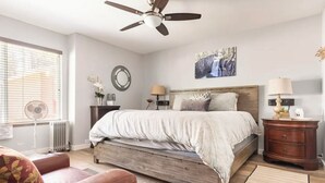 Ein Schlafzimmer mit einem Bett, Deckenventilator, Heizkörper, Fenster mit Jalousien, Wandspiegel und Nachttisch mit Lampe.