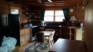 Cozinha compacta com armários de madeira, geladeira, fogão e micro-ondas. Há uma mesa de jantar com cadeiras, uma janela com cortinas e um relógio na parede.