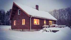 Uma cabana de madeira com telhado e janelas cobertas de neve, em meio a uma paisagem nevada.
