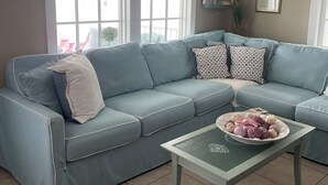 Un salón con un sofá azul, una mesa de centro de vidrio con objetos decorativos y ventanas amplias con persianas.