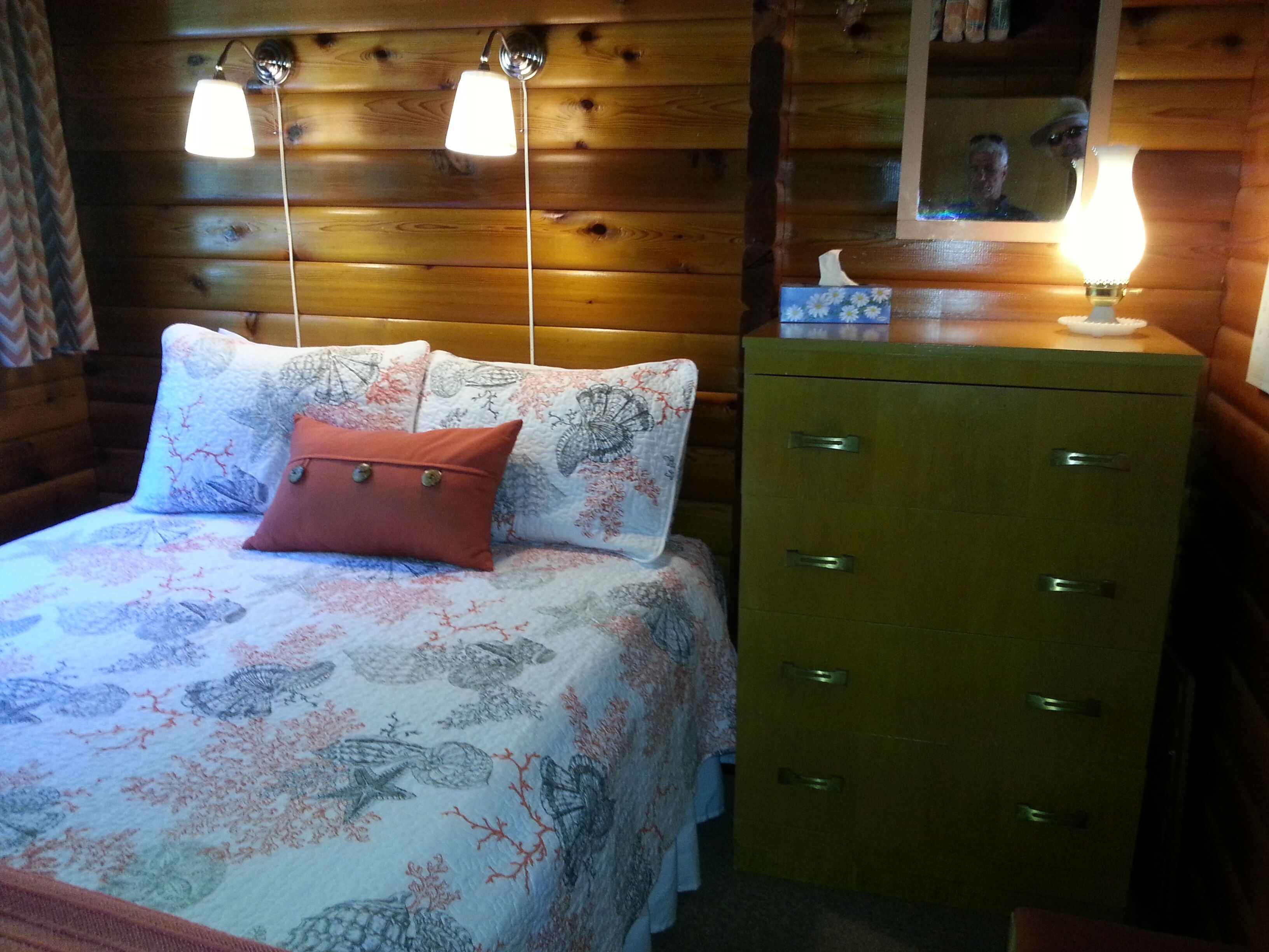 A wooden cabin room with a bed, two bedside lamps, a dresser, and a mirror on the wall.