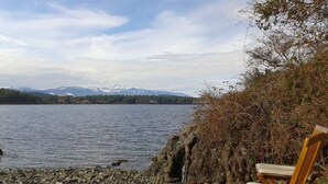 Una cadira de fusta a la riba rocosa d'un llac, amb vista a muntanyes nevades a la llunyança.