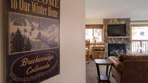 Una sala de estar con chimenea, un sofá y un comedor con mesa y sillas. Un letrero en la pared dice "Welcome to Our Winter Home Breckenridge Colorado".