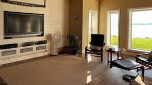 Een woonkamer met een flatscreen televisie, een ventilator, een stoel, een tafel en grote ramen met uitzicht op een gazon.