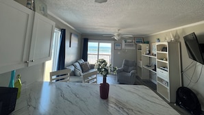Ein geräumiges Wohnzimmer mit einem großen Fenster zum Meer, einem Deckenventilator und einem Esstisch aus Marmor.