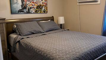 A bedroom with a bed, a nightstand, a lamp, an air conditioning unit, and a painting of a coastal town.