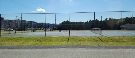 Cancha deportiva cercada, cielo despejado, edificios al fondo y una carretera en primer plano.