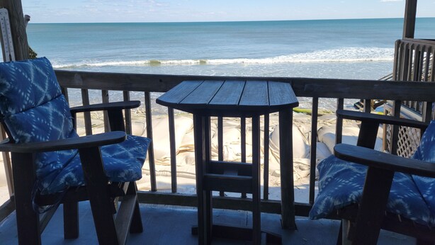 Eine Holzterrasse mit zwei Stühlen und einem Tisch mit Blick auf einen Strand und Wellen.