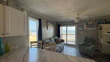 A spacious living area with a large window offering a view of the beach, a ceiling fan, and a dining area with a marble table.