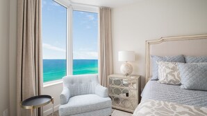 Un dormitorio con un ventanal grande, una silla, una mesita de noche con lámpara y una cama con ropa de cama estampada.