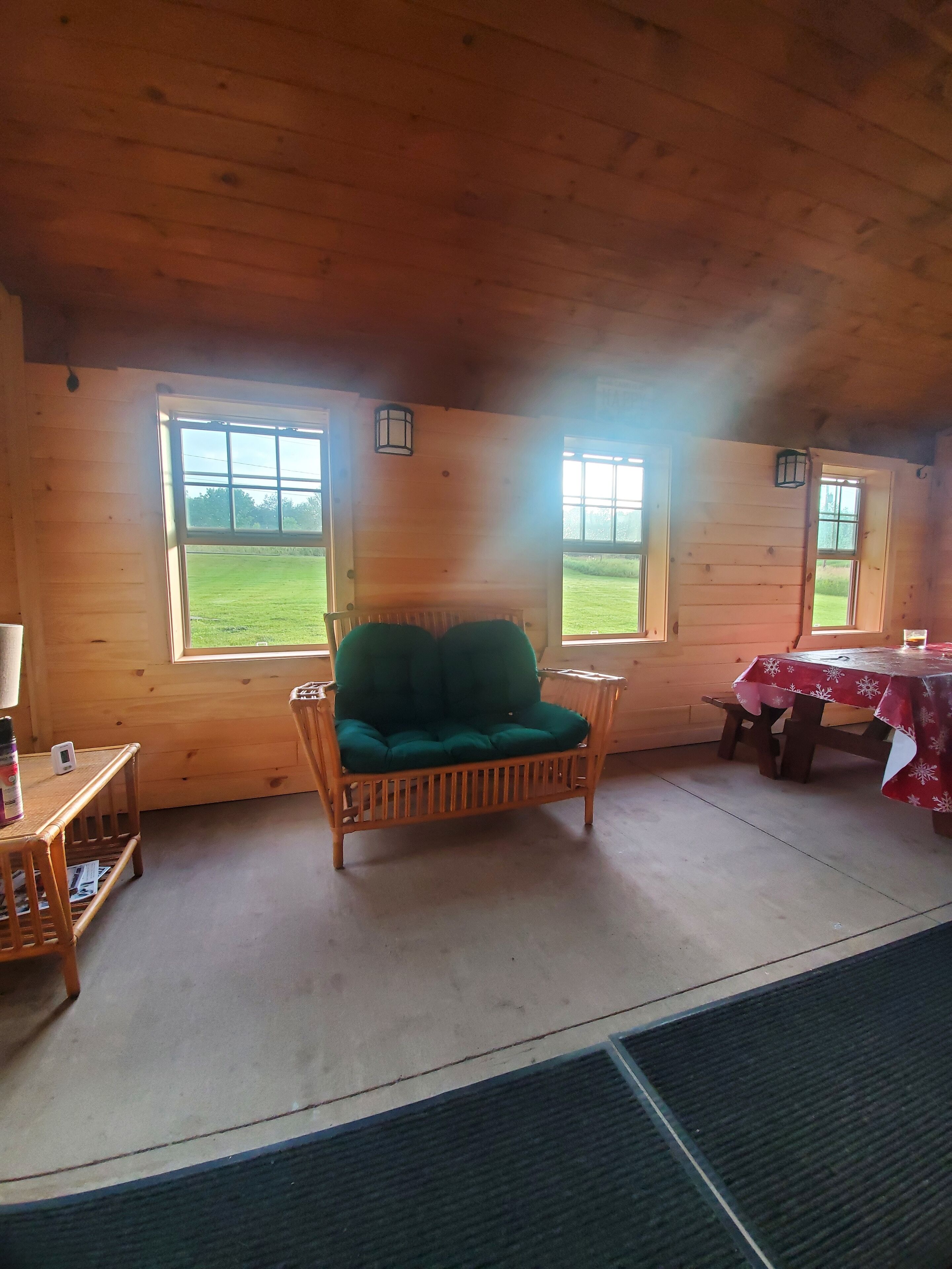 A wooden cabin interior with a green cushioned chair, a small table with a lamp, and a dining table with a red tablecloth.