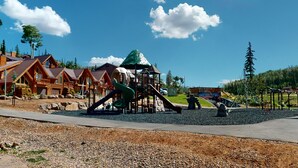A playground with a slide and climbing structures, surrounded by wooden cabins and a clear sky.