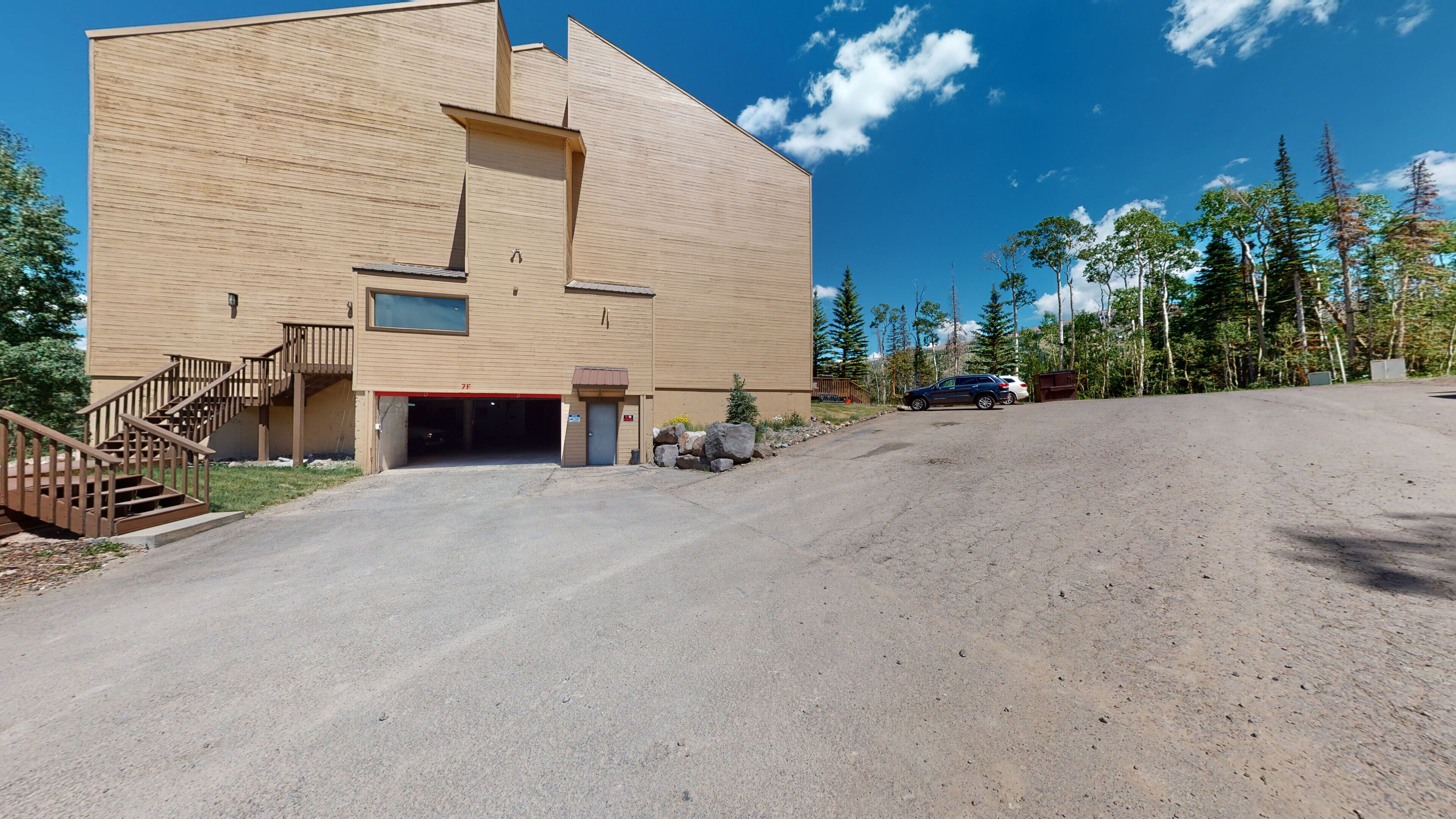 A modern building with a garage entrance, a staircase, and a dirt driveway leading to parked cars.