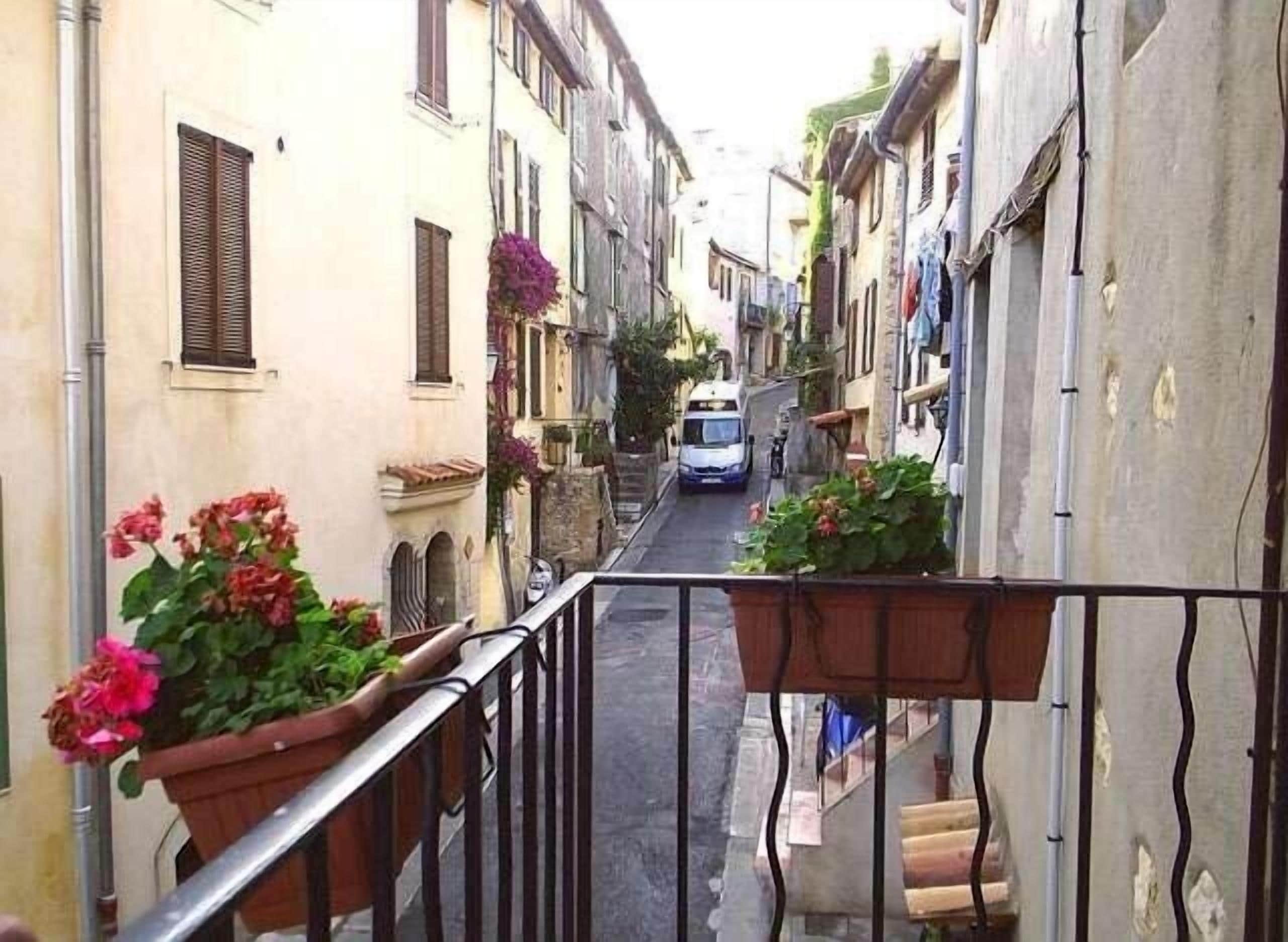 Une rue étroite, bordée de bâtiments des deux côtés, avec un fourgon blanc garé au milieu, et des plantes en pot, fleuries, sur les balcons.
