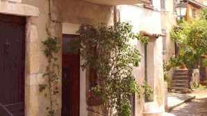A narrow street with a stone building featuring a dark wooden door and a small garden area with a potted plant and a red flower. - Charming Village House In Medieval Village, Close To Beach And Nice (Cagnes-Sur-Mer)