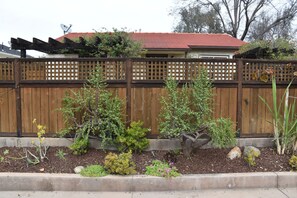 Una valla de madera con diseño de celosía, un jardín con diversas plantas y un árbol pequeño, y un edificio con techo rojo al fondo.