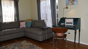 Una sala de estar con un sofá seccional, una mesita, un sillón, un escritorio con una lámpara y un cuadro en la pared.