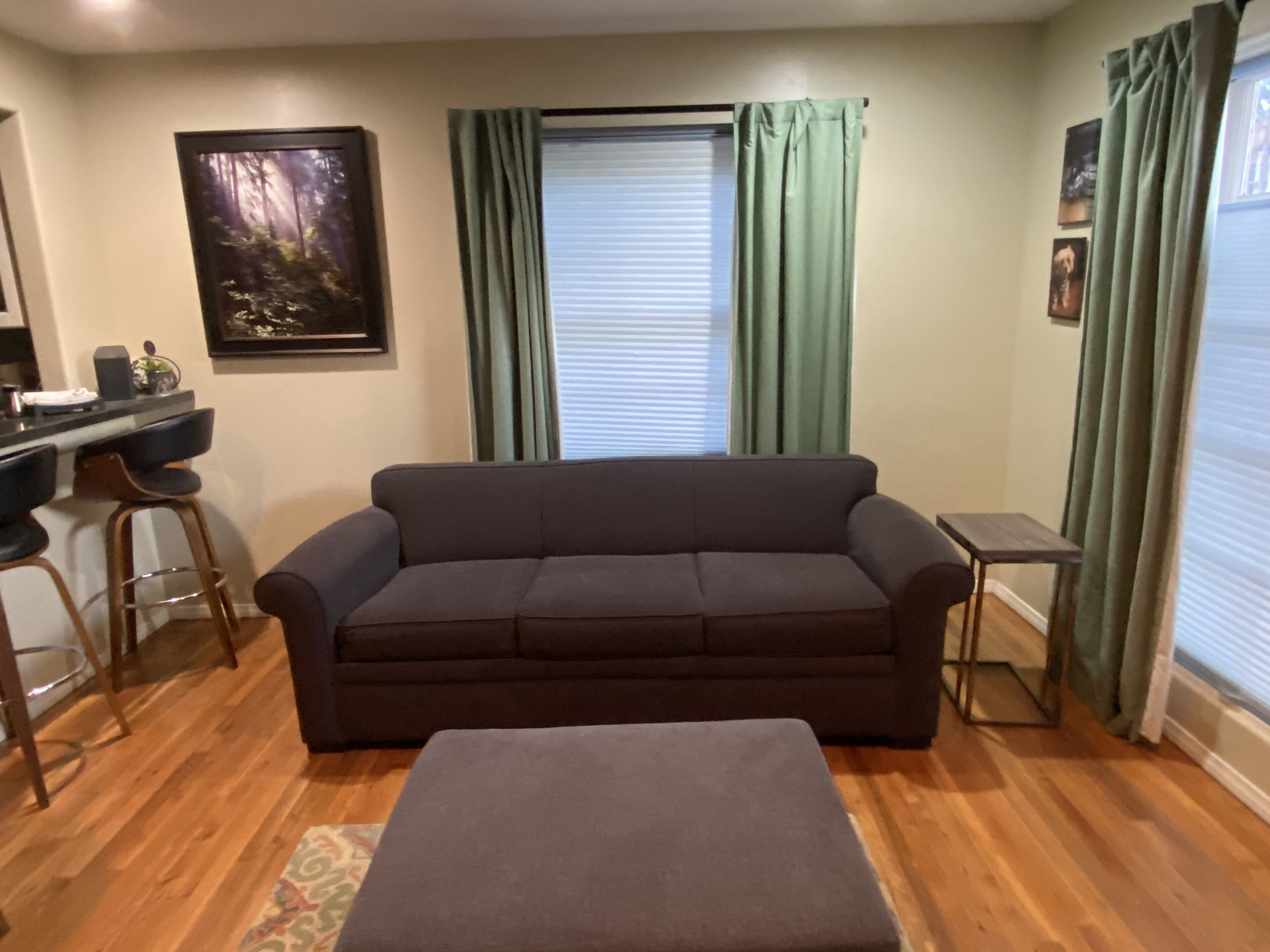 A living room with a sofa, a small table, a bar stool, a window with curtains, and a framed picture on the wall.